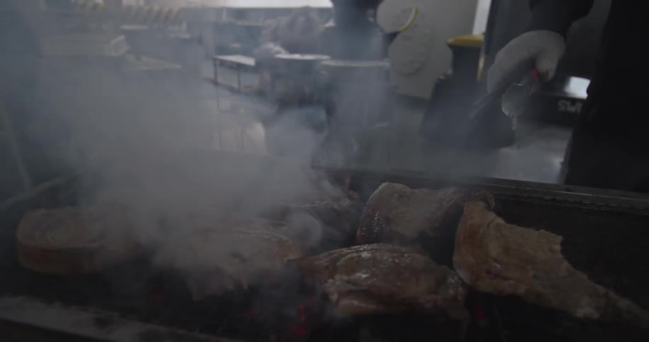 Thick cuts of meat sizzle on a smoky barbecue grill in an industrial setting in slow motion