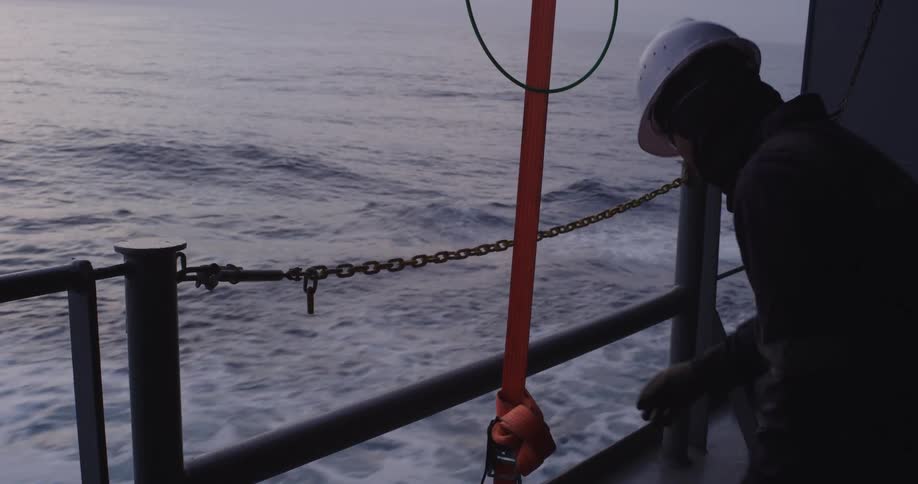 A crew member in a hard hat and protective gear works on the deck of a ship sailing through the ocean at twilight
