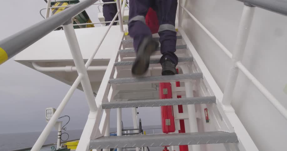 Slow motion shot of a crew member in work boots and coveralls walking up the metal stairs on the deck of a ship