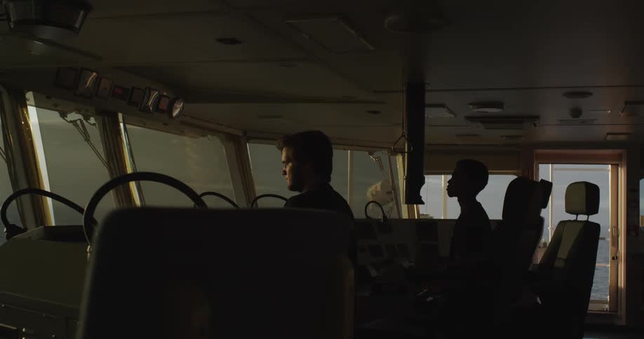 Two silhouetted crew members navigate a large vessel from the ship's bridge as the golden sun rises over the ocean