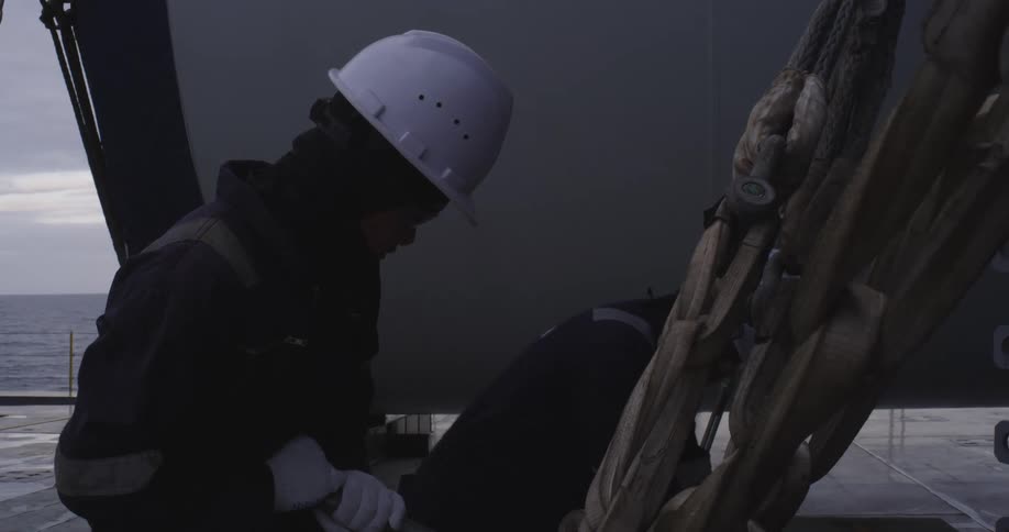 Two deckhands in hard hats and workwear meticulously handle thick ropes on the deck of a ship at sea