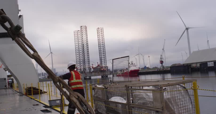 A dock worker in safety gear secures rigging on a ship's deck in an industrial harbor with wind turbines and an oil rig in the background