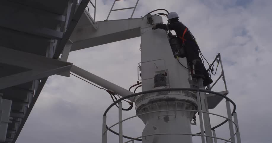 From a low angle, a worker wearing a safety harness and hard hat works on a ship's crane against an overcast sky