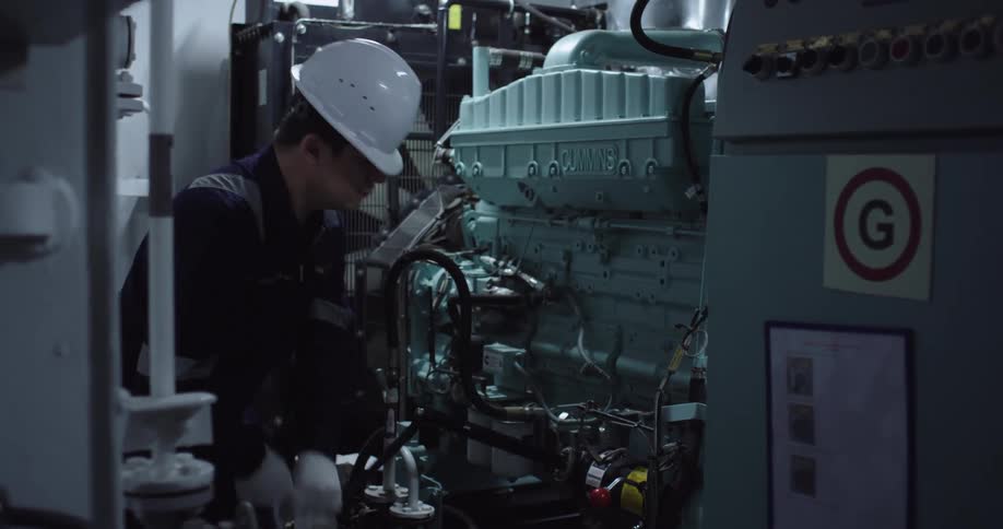 A marine engineer in a hard hat services a large Cummins diesel generator inside a ship's engine room