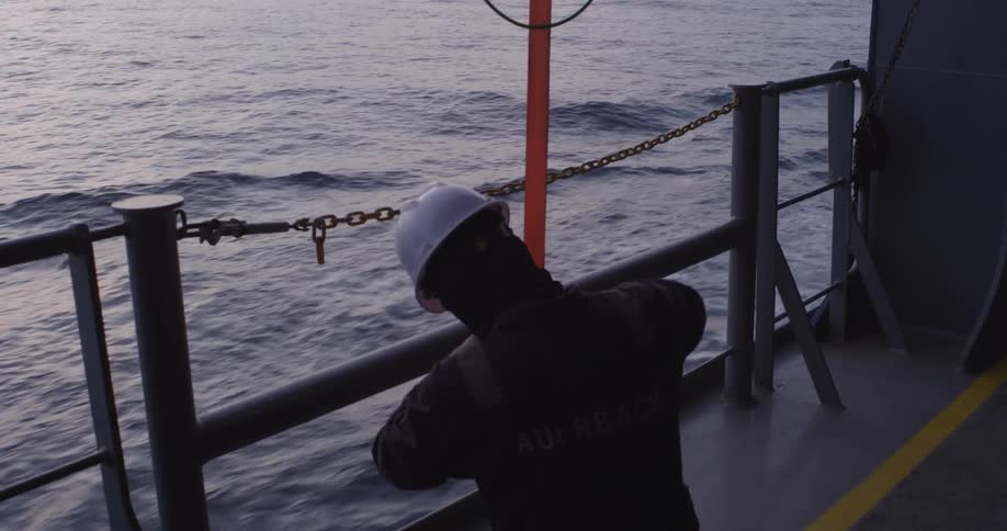 A ship's crew member in a hard hat uses a ratchet strap to secure a post on deck while sailing on the ocean at dusk
