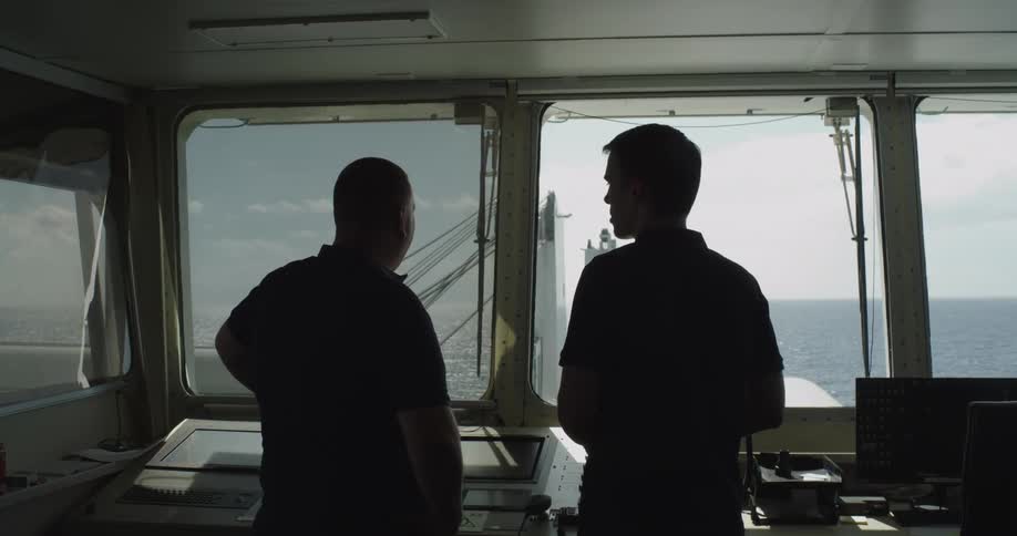 Two silhouetted crew members chat on the bridge of a cargo ship as it sails across the open ocean