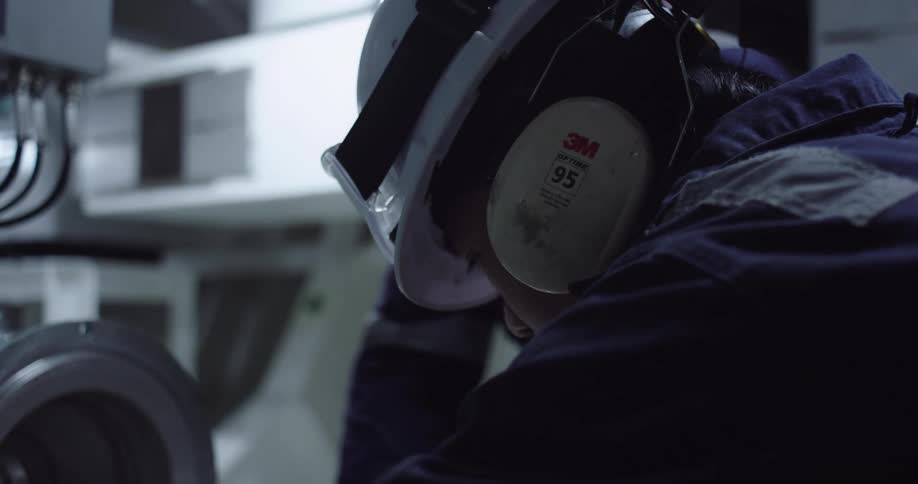An industrial worker wearing a helmet and earmuffs diligently performs maintenance on machinery in a factory setting