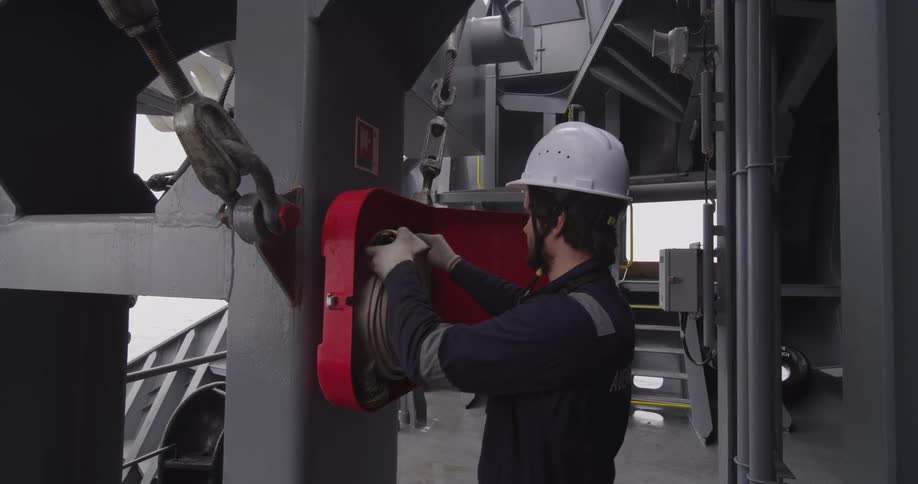 A crew member in a hard hat and protective gear carefully connects a nozzle to a fire hose on a large ship