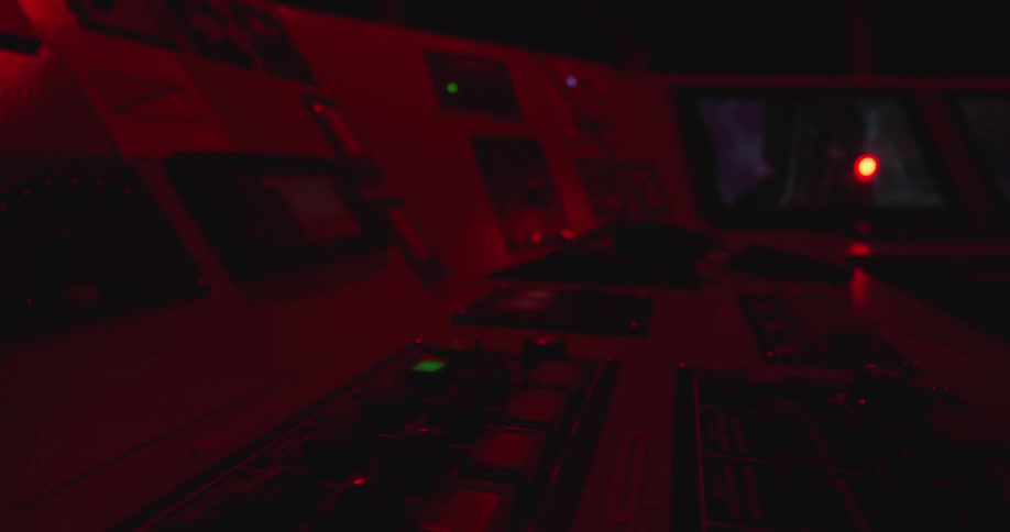 An out-of-focus control panel in a dark command center is illuminated by blinking red emergency lights