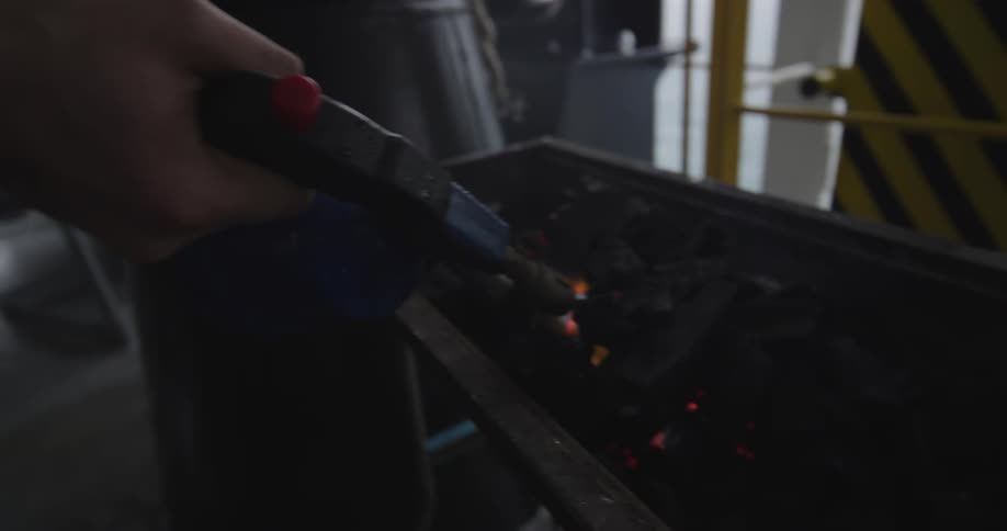 A worker carefully lights charcoal with a blowtorch and begins to heat a blue industrial part in a workshop