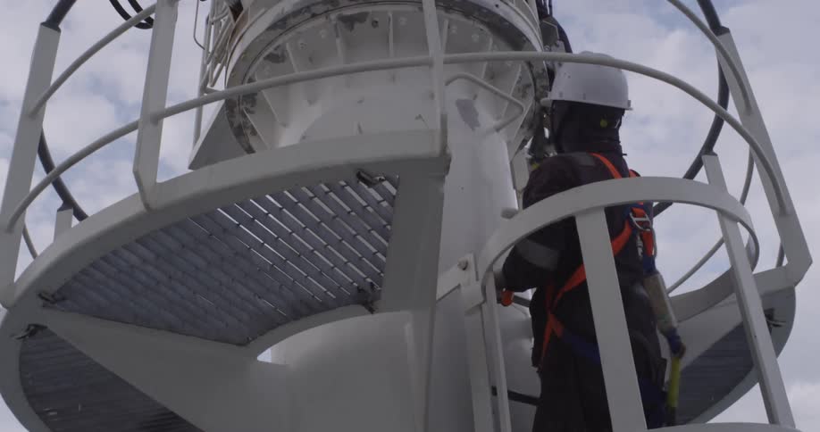 Low angle shot of an industrial painter in full safety gear working at height, painting a large white metal structure against an overcast sky