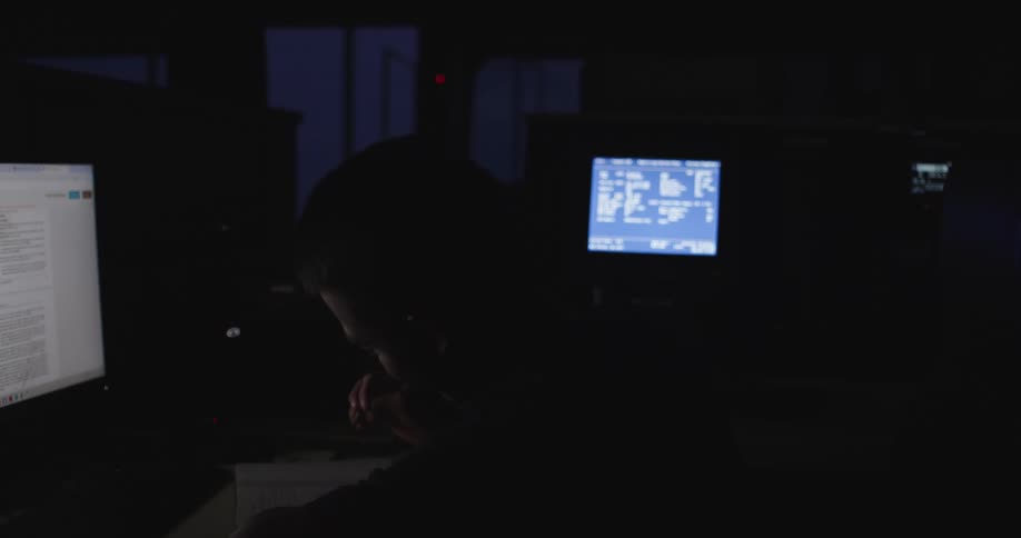 A focused person takes notes in a notebook while sitting in a dark room surrounded by glowing computer screens
