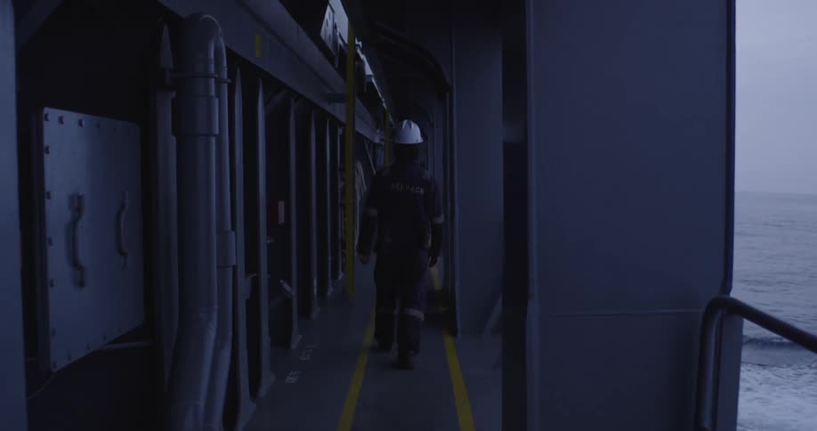 A lone seafarer wearing a hard hat walks slowly down a dimly lit exterior corridor of a large vessel sailing on the open ocean
