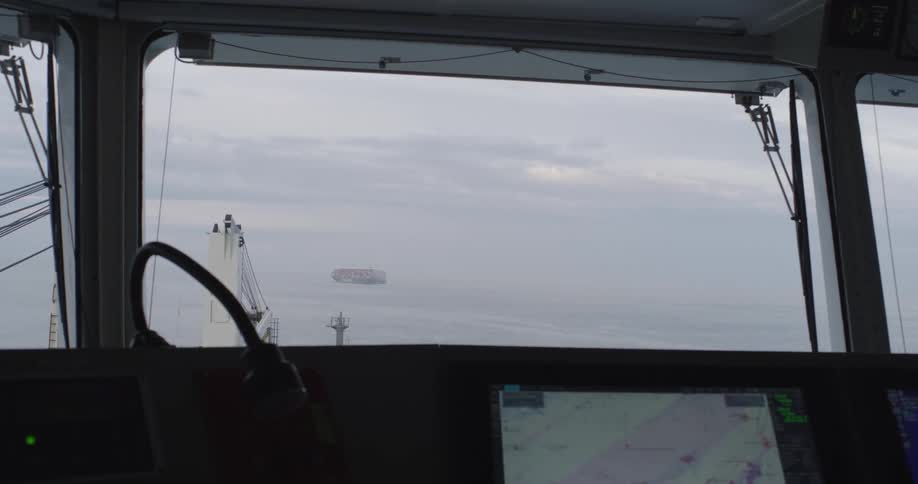 A large container ship sails on the hazy open sea, as seen from the navigation bridge of another vessel