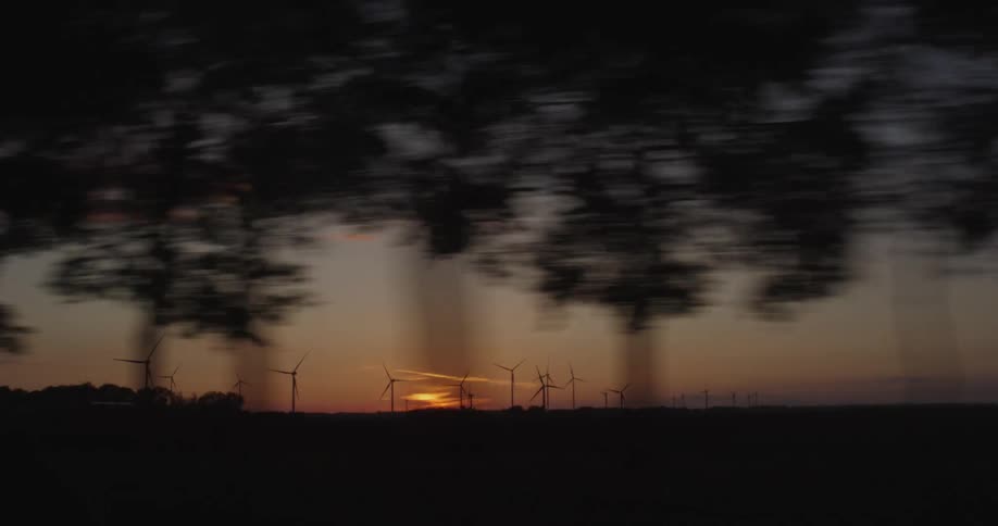 A dreamy driving shot captures silhouetted wind turbines against a vibrant sunset sky, with blurred trees passing in the foreground