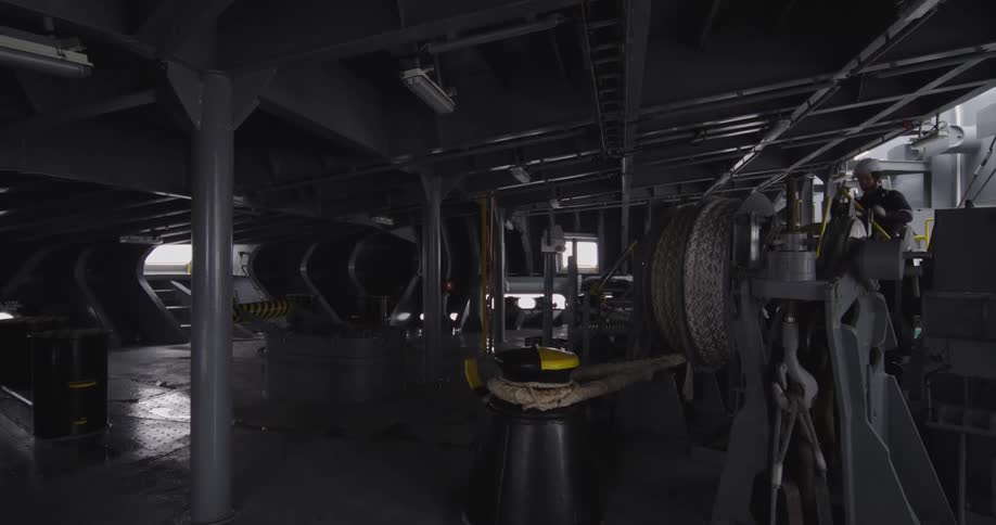A seaman in a hard hat operates a large mooring winch on the industrial deck of a vessel