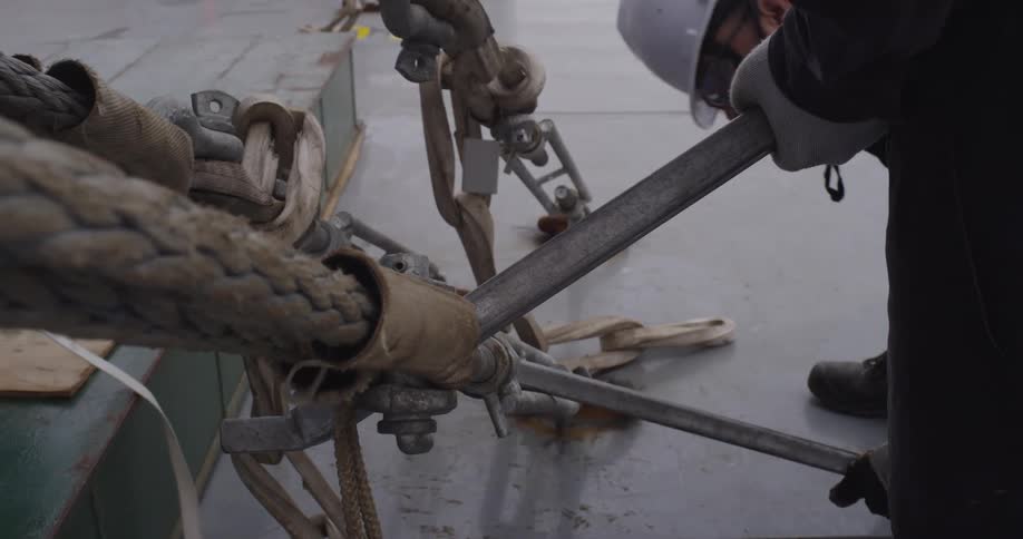 Close up of industrial workers using levers and heavy-duty ropes to lash down equipment on a ship's deck