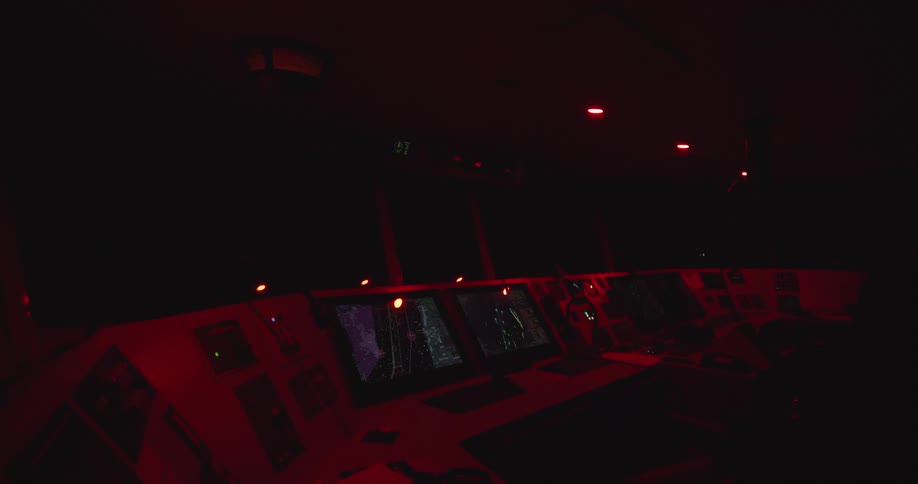 A ship's navigation bridge is bathed in the red glow of its control panels and screens during a nighttime operation