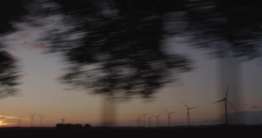 A tranquil tracking shot from a moving vehicle captures a sprawling wind farm with turbines silhouetted against the vibrant colors of a setting sun