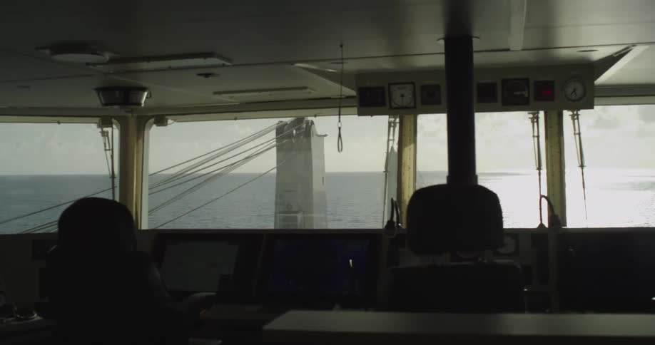 A slow pan across the dimly lit bridge of a commercial ship sailing across the open ocean toward the horizon