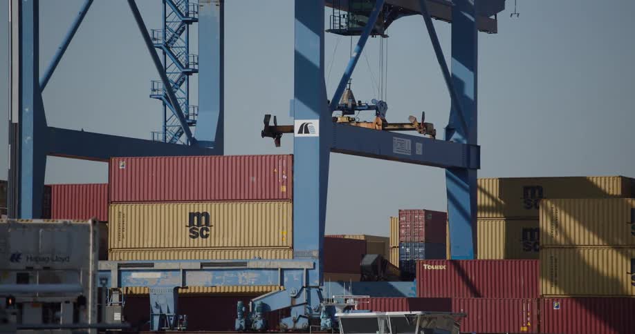 A large blue gantry crane moves its spreader across a yard full of stacked shipping containers at an industrial port