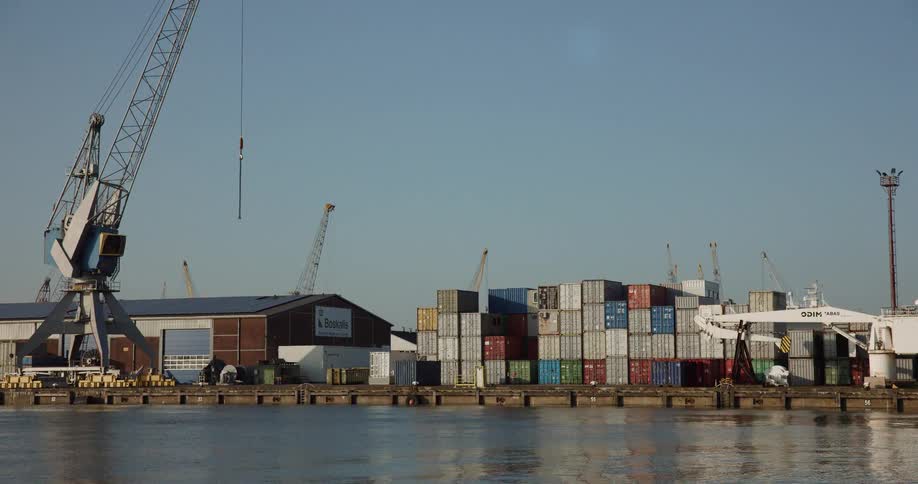 A static wide shot of a busy industrial harbor shows colorful shipping containers stacked on the dock next to large cranes