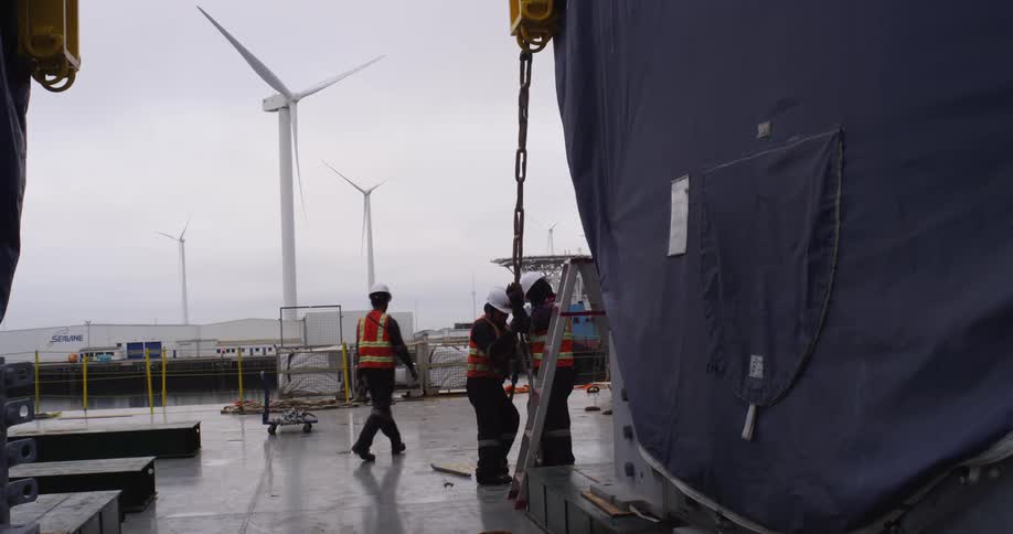 Industrial workers in safety gear secure heavy cargo with chains on a ship deck with wind turbines in the background