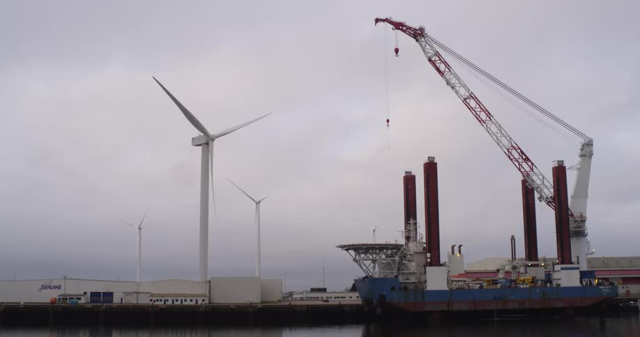 An offshore wind turbine installation vessel with a massive crane is docked in a harbor next to spinning wind turbines on a cloudy day