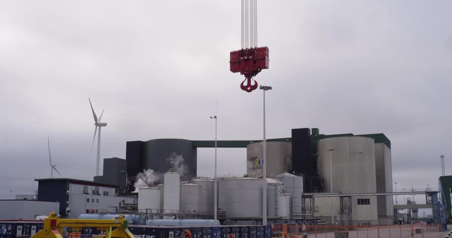 A static shot of an industrial plant with steaming silos and a large red crane hook, while wind turbines spin in the background under an overcast sky