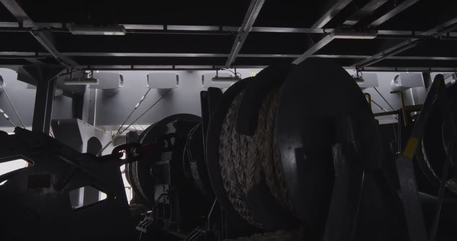 A heavy-duty industrial winch slowly pulls in a thick rope and chain in a dark engine room