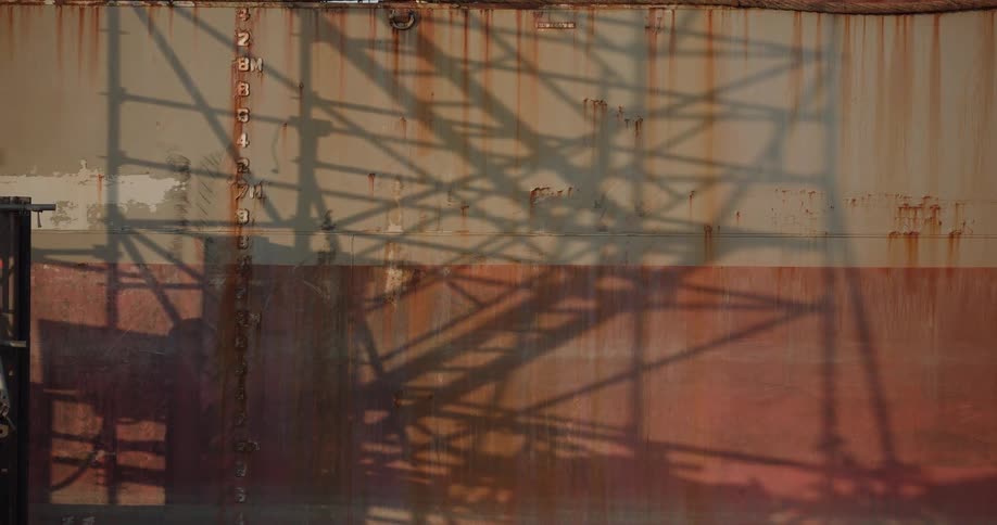The shadow of an industrial crane is cast upon the rusty hull of a cargo ship at port