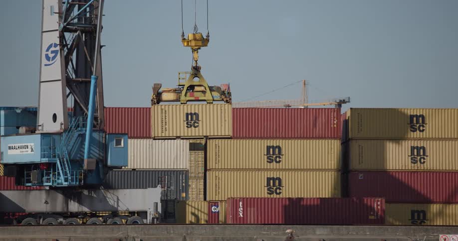 A gantry crane carefully lifts and stacks a large cargo container in an industrial shipping yard on a sunny day
