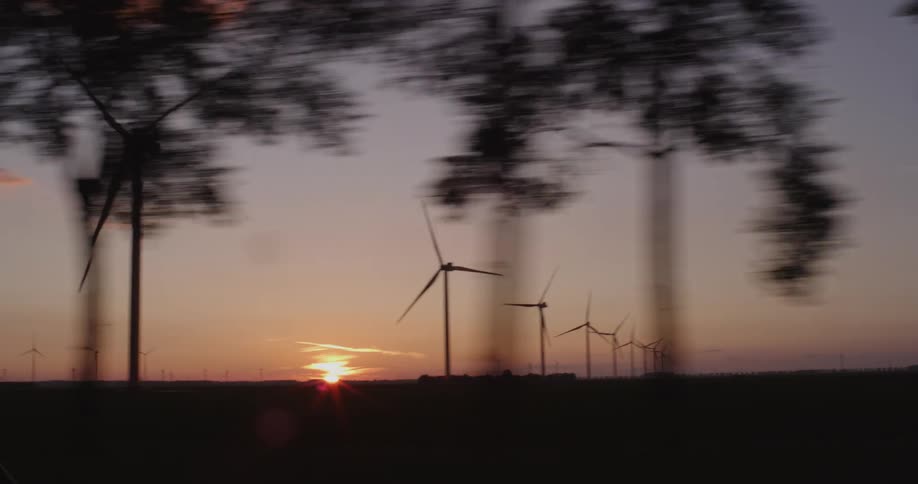 Driving past silhouetted wind turbines on a rural wind farm as the sun sets