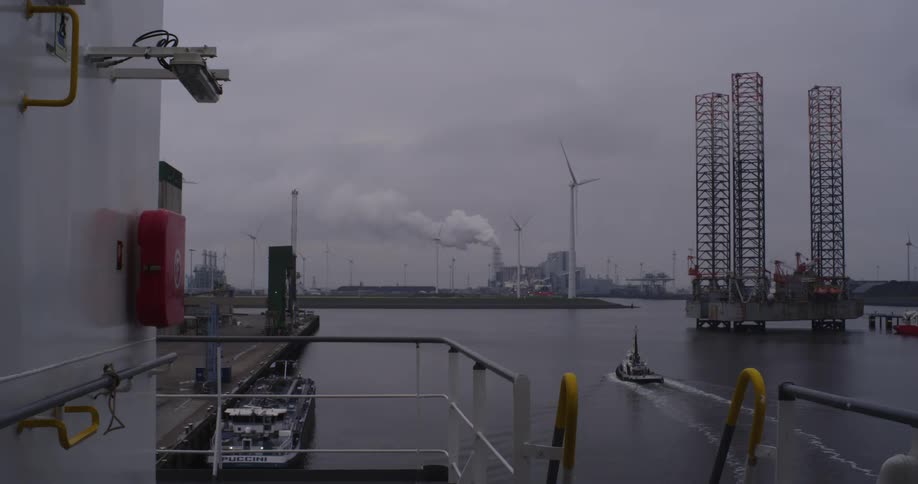 A view from a ship shows a large industrial harbor with an offshore oil rig, wind turbines, a power plant, and a tugboat sailing on a cloudy day