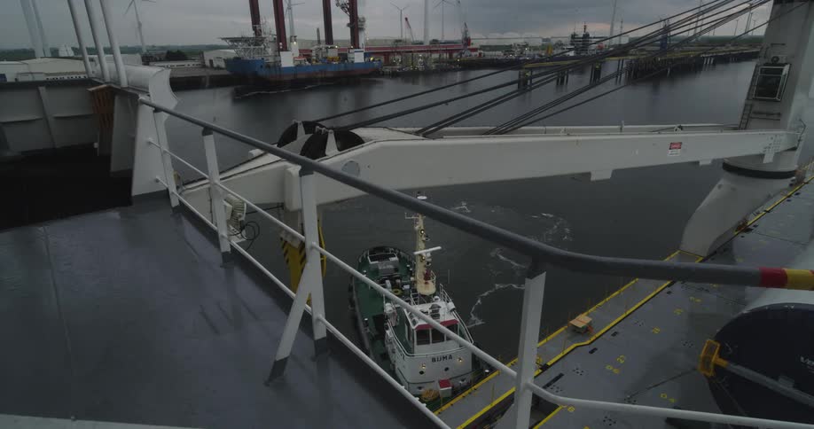 A tugboat idles alongside a large ship in an industrial port on a cloudy, overcast day