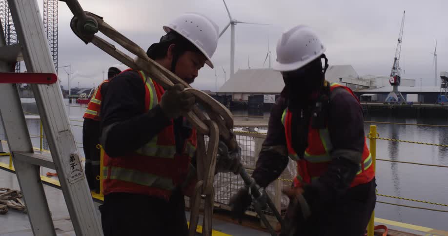 Construction workers in hard hats prepare heavy chains and rigging on a vessel docked at an industrial port with wind turbines
