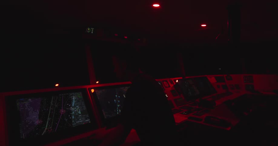 A ship's navigation officer monitors radar and electronic charts on the bridge during a night watch, with the control room bathed in red light
