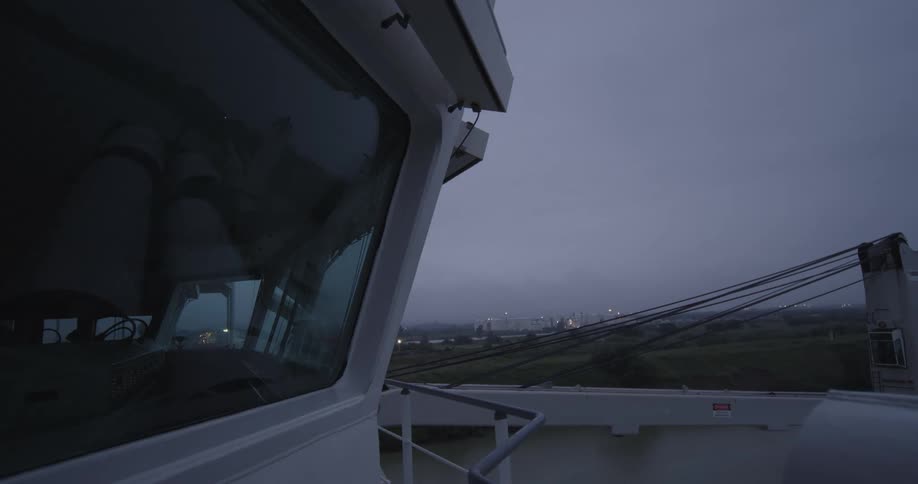 A moody, static shot from the deck of a large vessel looking towards a distant industrial area on a gloomy, overcast day
