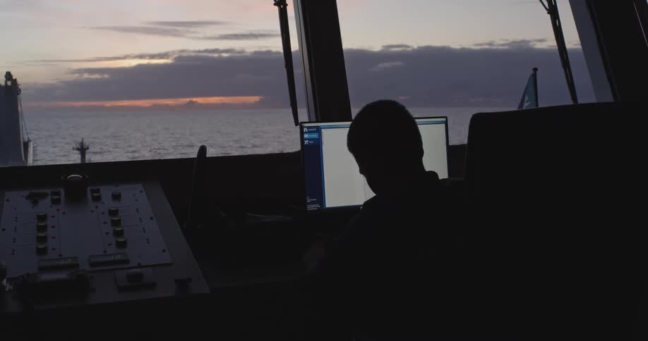 A silhouetted figure works at a computer console on the bridge of a ship, with the ocean and a sunset visible through the window