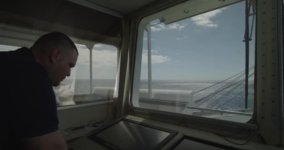 A sailor navigates from the ship's bridge, with a clear view of the sunlit open ocean ahead