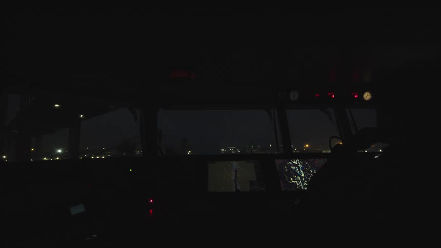 A pilot sits in the dark cockpit of a plane at night, looking out at the lights of the airfield