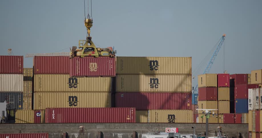 An industrial crane lifts and moves a red cargo container, stacking it at a busy shipping terminal