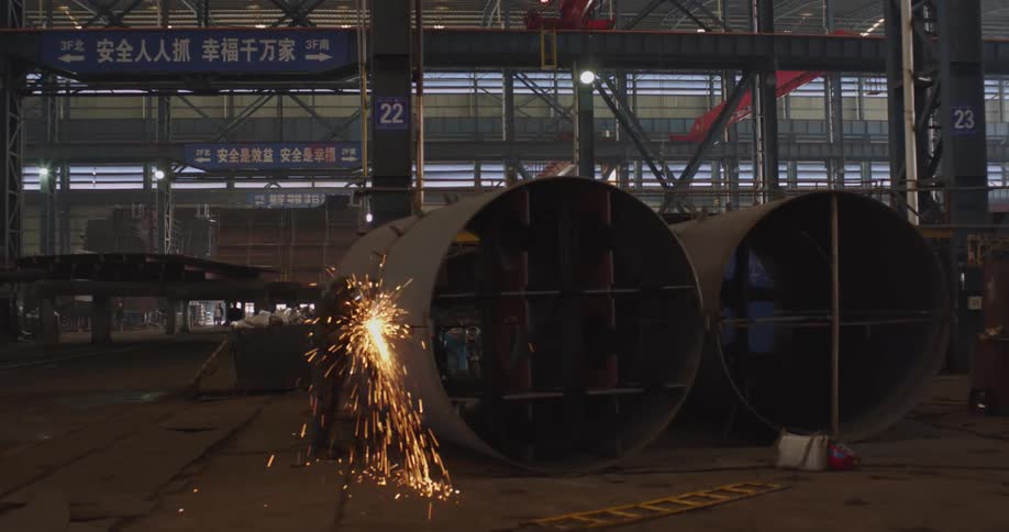 Sparks illuminate a dark factory as a worker grinds a large metal cylinder in an industrial setting