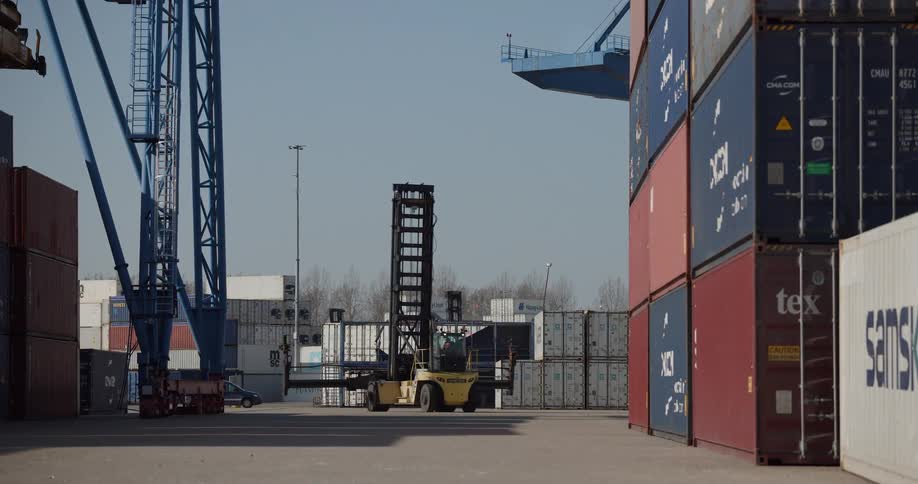 A yellow reach stacker maneuvers between stacks of shipping containers at an industrial port terminal on a sunny day