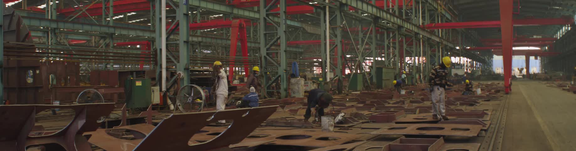 Workers in a massive industrial shipyard use tools to weld large metal plates, creating bright sparks