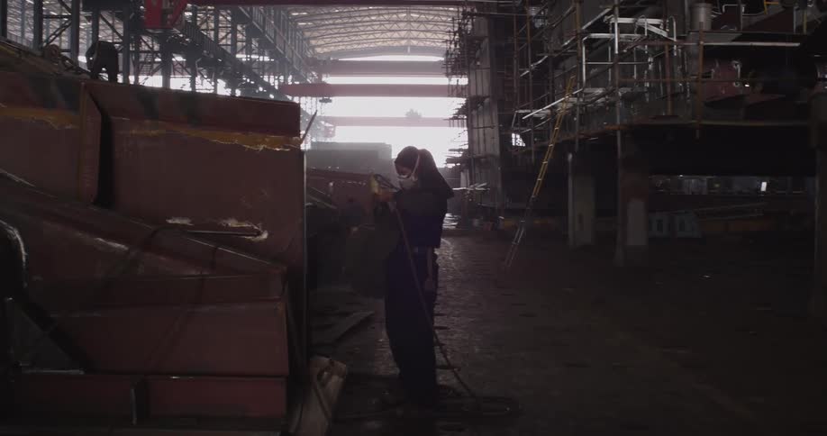 A worker in a respirator mask uses an angle grinder on a metal structure in a vast, dark industrial workshop