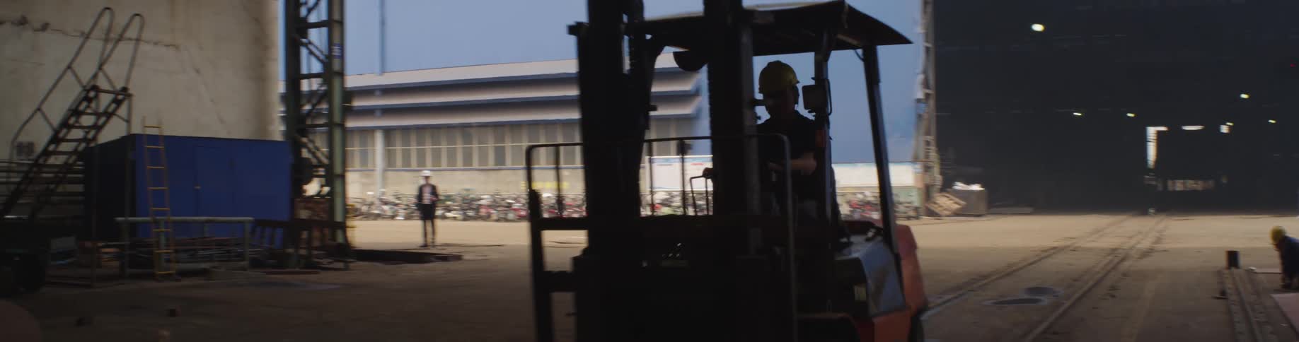 A factory worker in a hard hat drives a forklift from a bright industrial lot into a dark warehouse