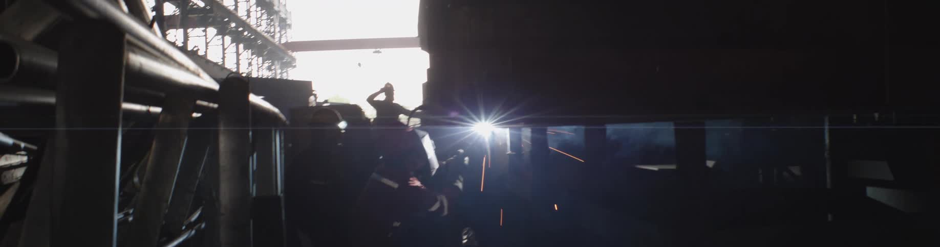 A welder works in a dark factory, the bright light from their torch creating a lens flare as sparks fly