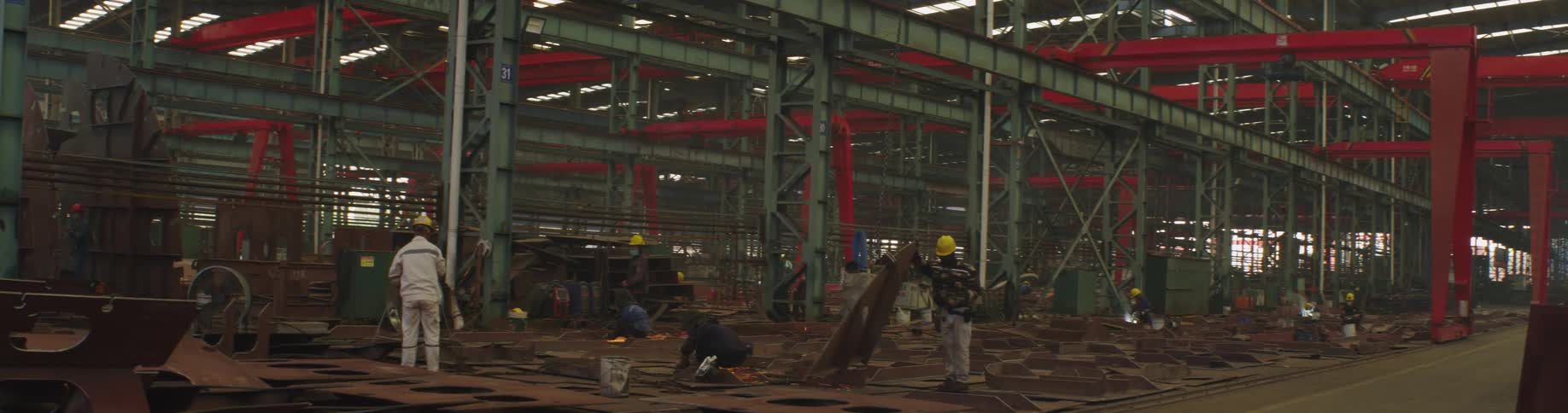 Workers grind and weld large steel plates on the floor of a bustling industrial shipyard factory