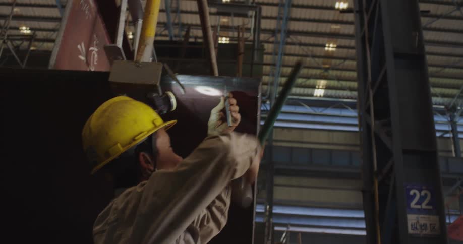 A worker in a yellow hard hat meticulously inspects a weld on a large metal structure inside a manufacturing facility